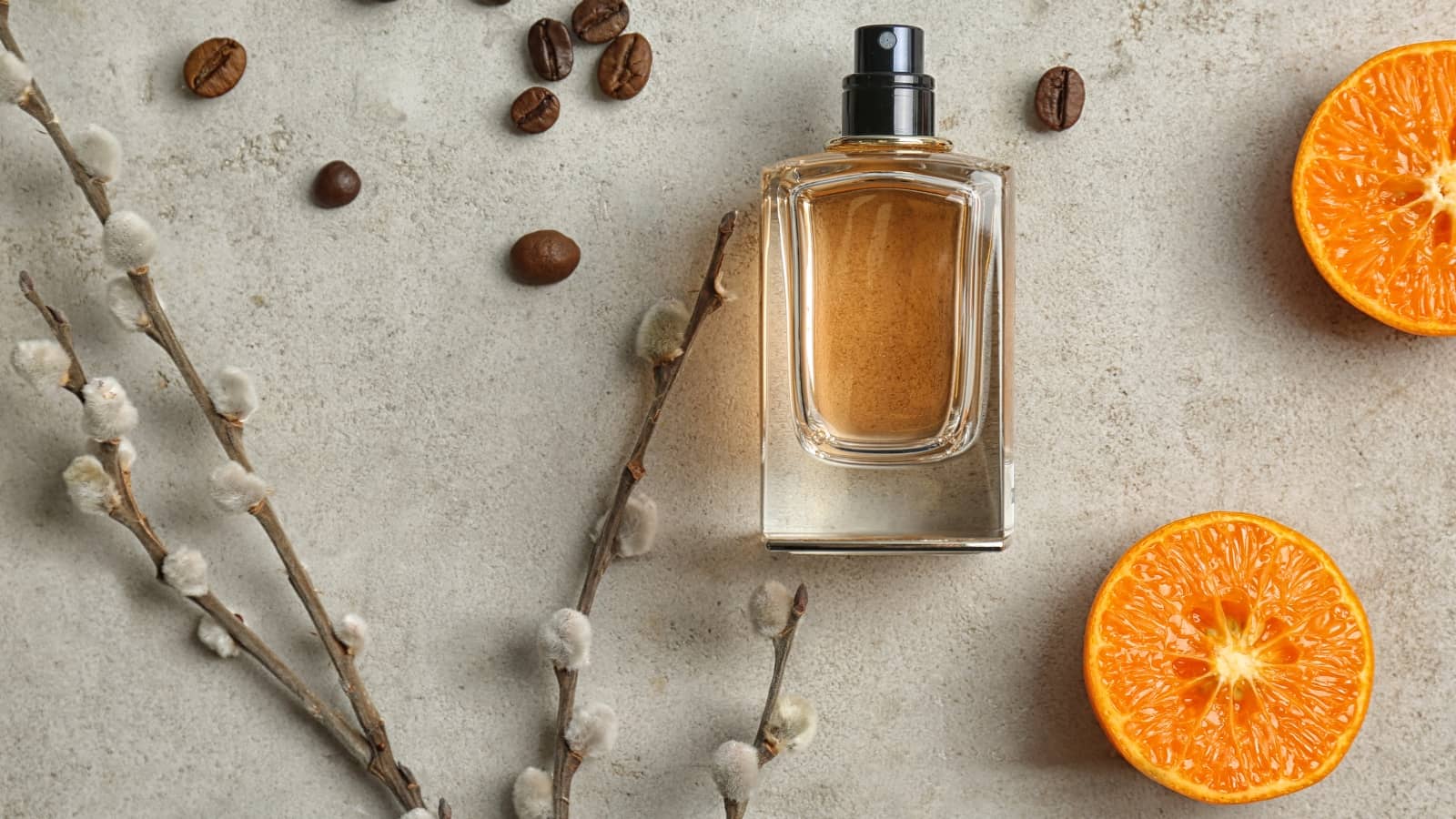 Maskuliner Flakon mit Kaffeebohnen, Osterzweig und Orangenscheiben vor grauem Hintergrund